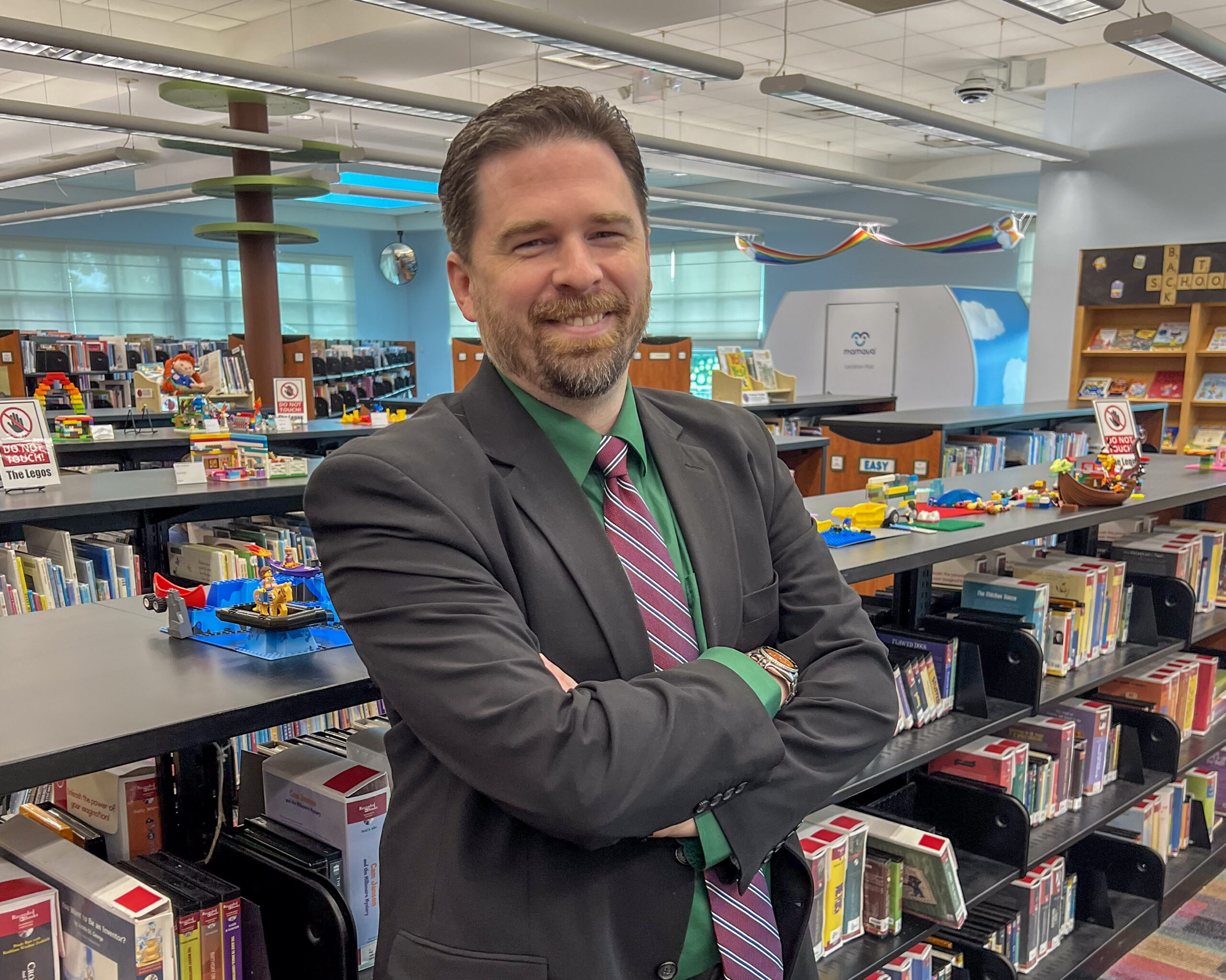 Library Director Dustin Goforth, a whilte man with brown hair and a grey suit, stands smiling in the Children's Department of the Blount County Public Library with colorful books and Lego projects in background