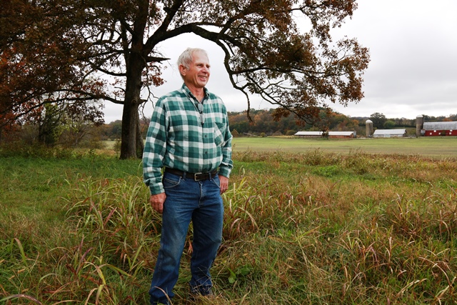 A white man with grey hair wearing a plaid shirt and jeans stands in a green field with a large tree behind him.