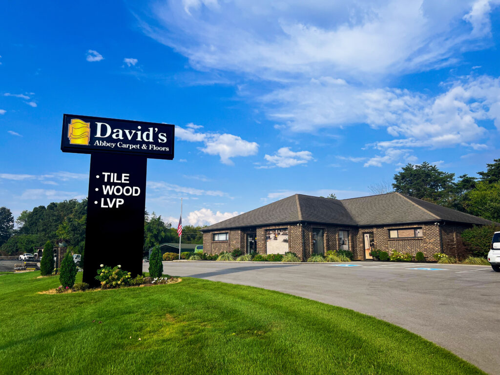 A brick building with a brown roof under a blue, partly clouded sky. A large black sign to the left reads David's Abbey Carpets and Floors, Tile, Wood, LVP.