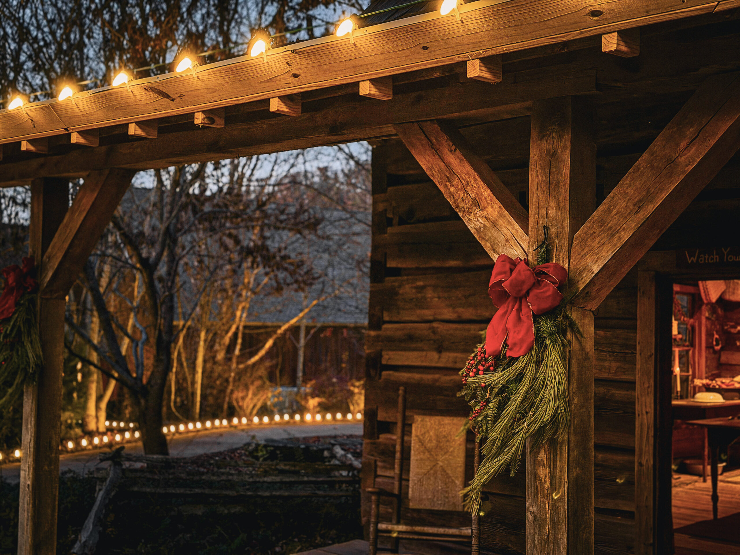 A rustic cabin porch with white holiday lights along the roof and a spray of pine branches with a red ribbon on one post.