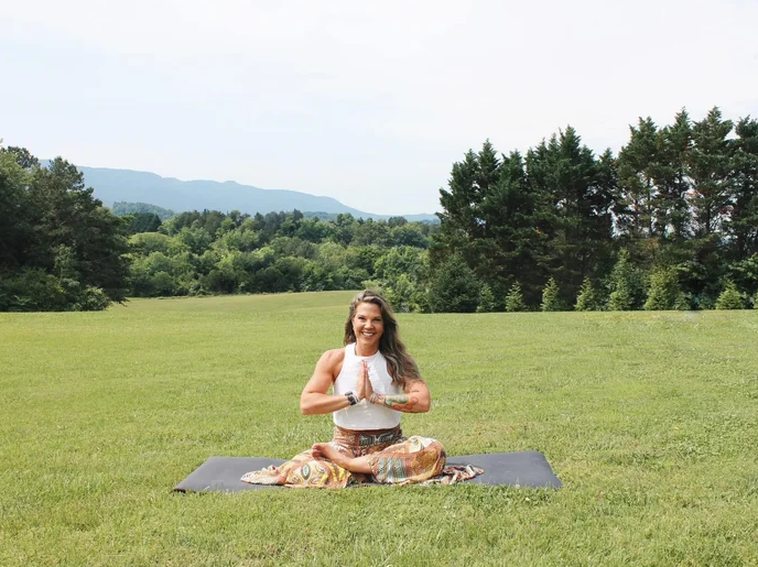 A white woman sits in full lotus position on a broad green lawn with rolling hills and trees in the background